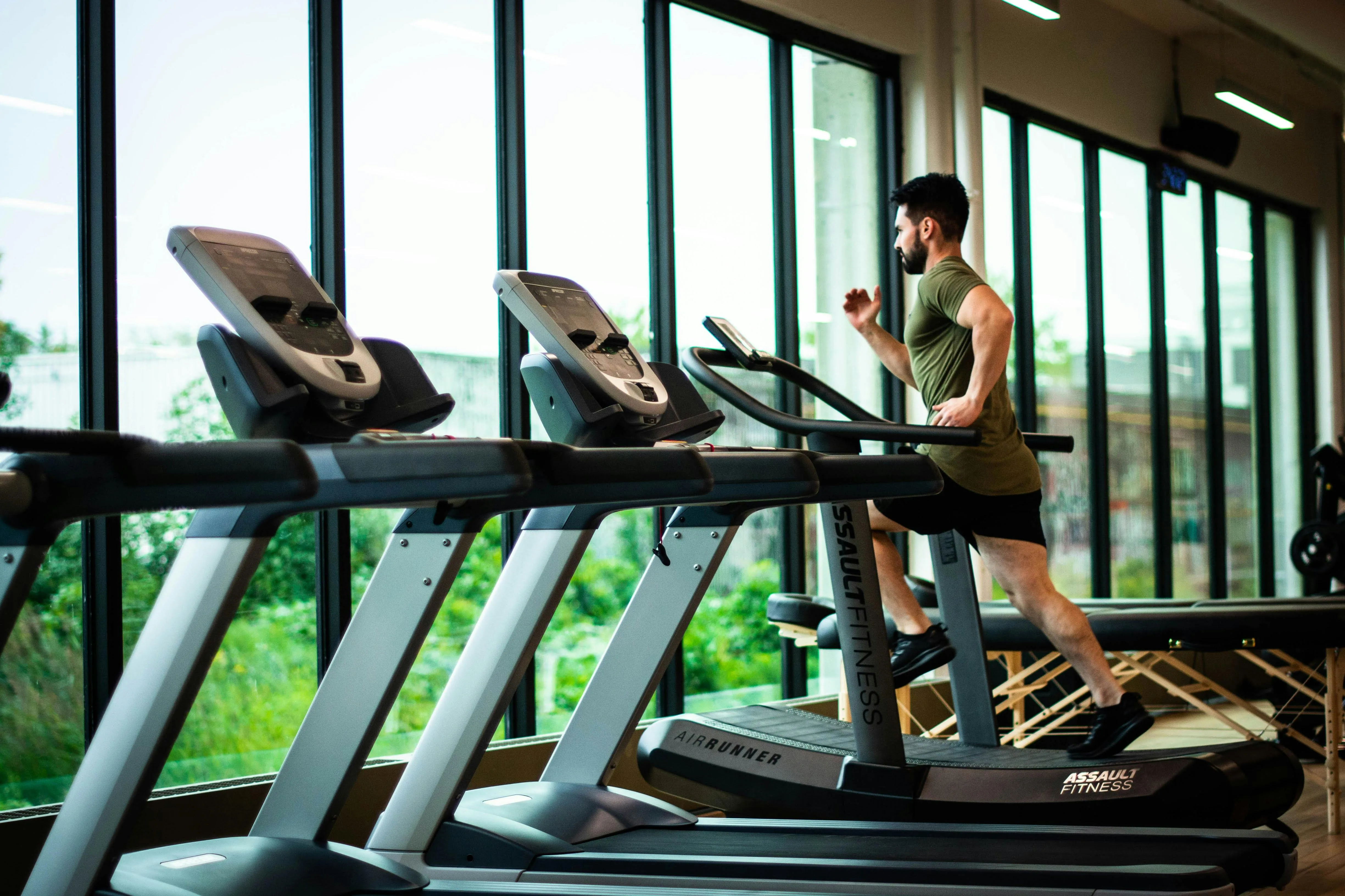 A man runs on a treadmill in a modern gym lined with large windows.